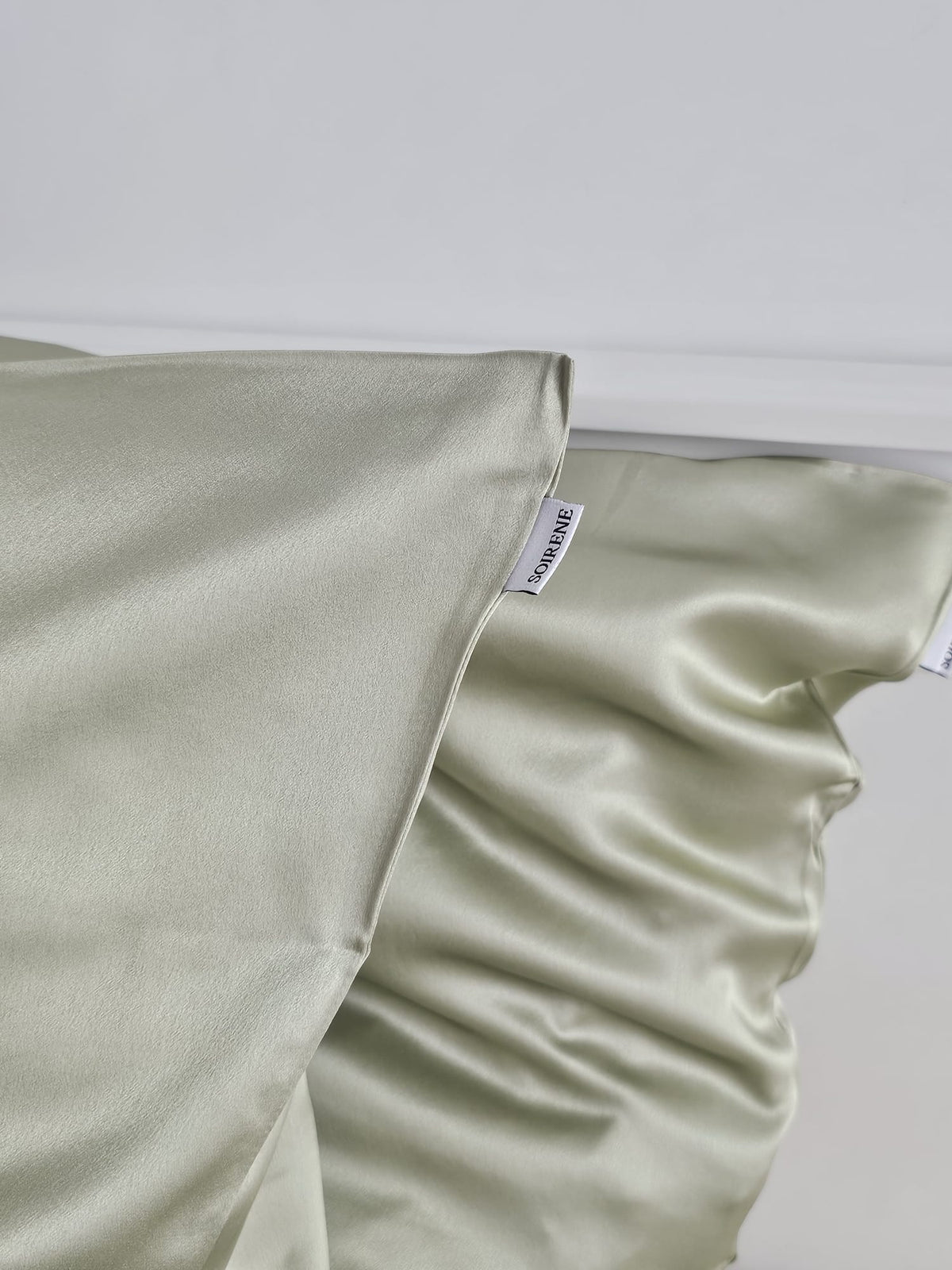 Gros plan sur une taie d'oreiller en soie de mûrier vert pistache SOIRENE montrant la couture soignée et l'étiquette de marque visible.