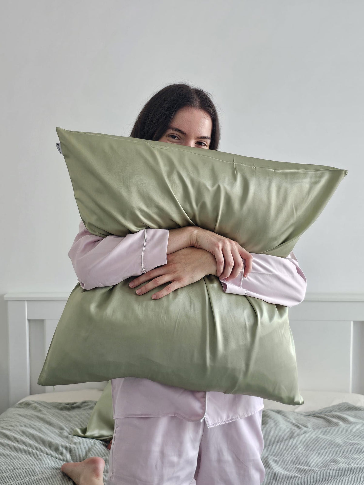 Femme souriante en pyjama rose tenant contre elle une taie d'oreiller en soie de mûrier vert pistache SOIRENE